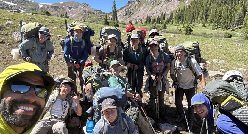 Instructors and students wearing backpacks pose for a group photo in a green alpine meadow 